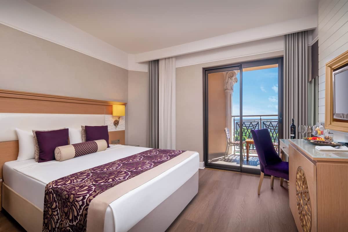 A hotel room with a large bed, a desk with a chair, a TV, and a balcony with a view.