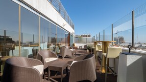 Terraço no telhado com mobiliário de vime e paredes de vidro, proporcionando vista da cidade.