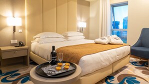 Quarto de hotel com uma cama, abajures, um tabuleiro com pastéis e uma garrafa, e vista para prédios pela janela.
