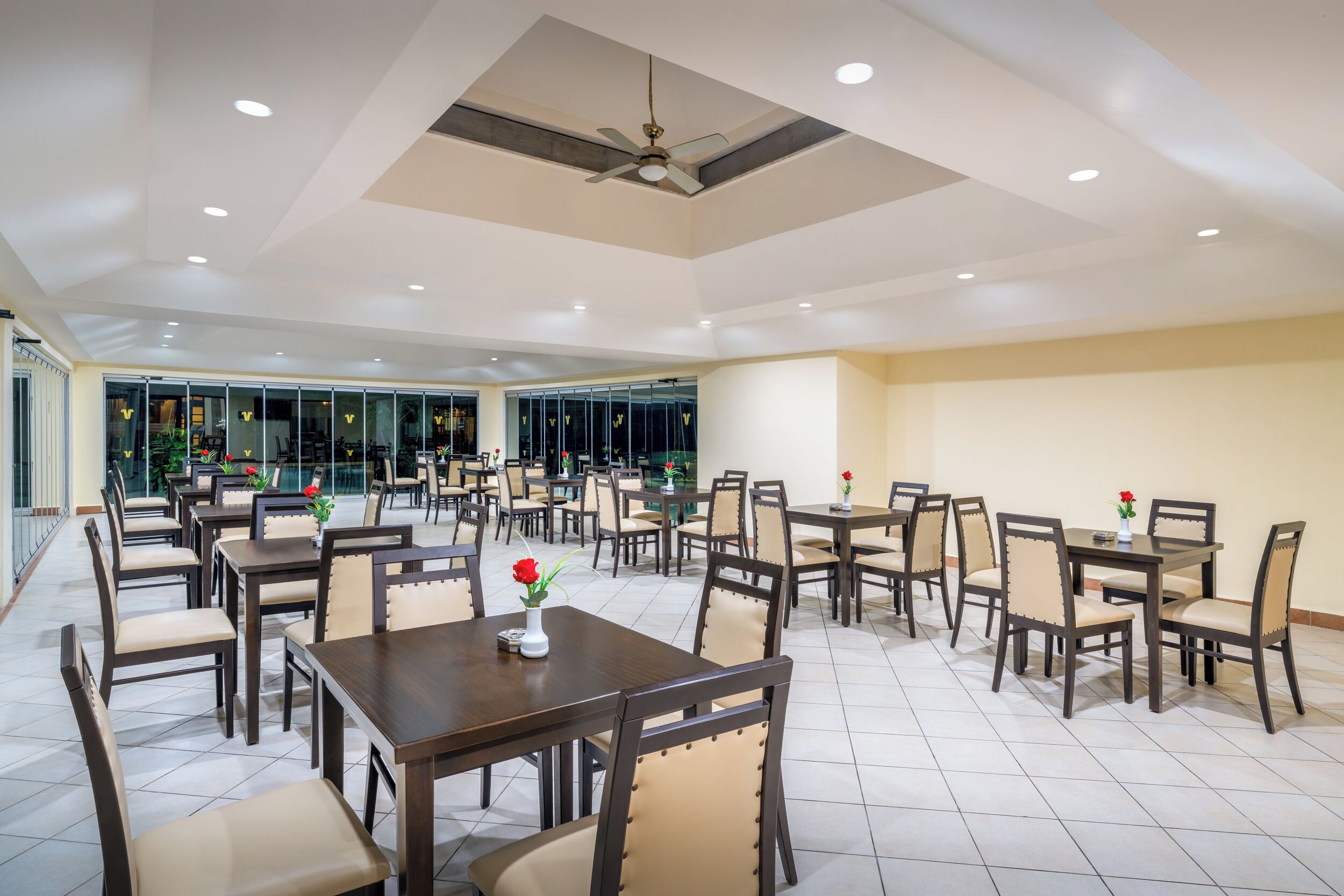 A spacious dining area with multiple tables and chairs, a ceiling fan, and a view through large glass windows.