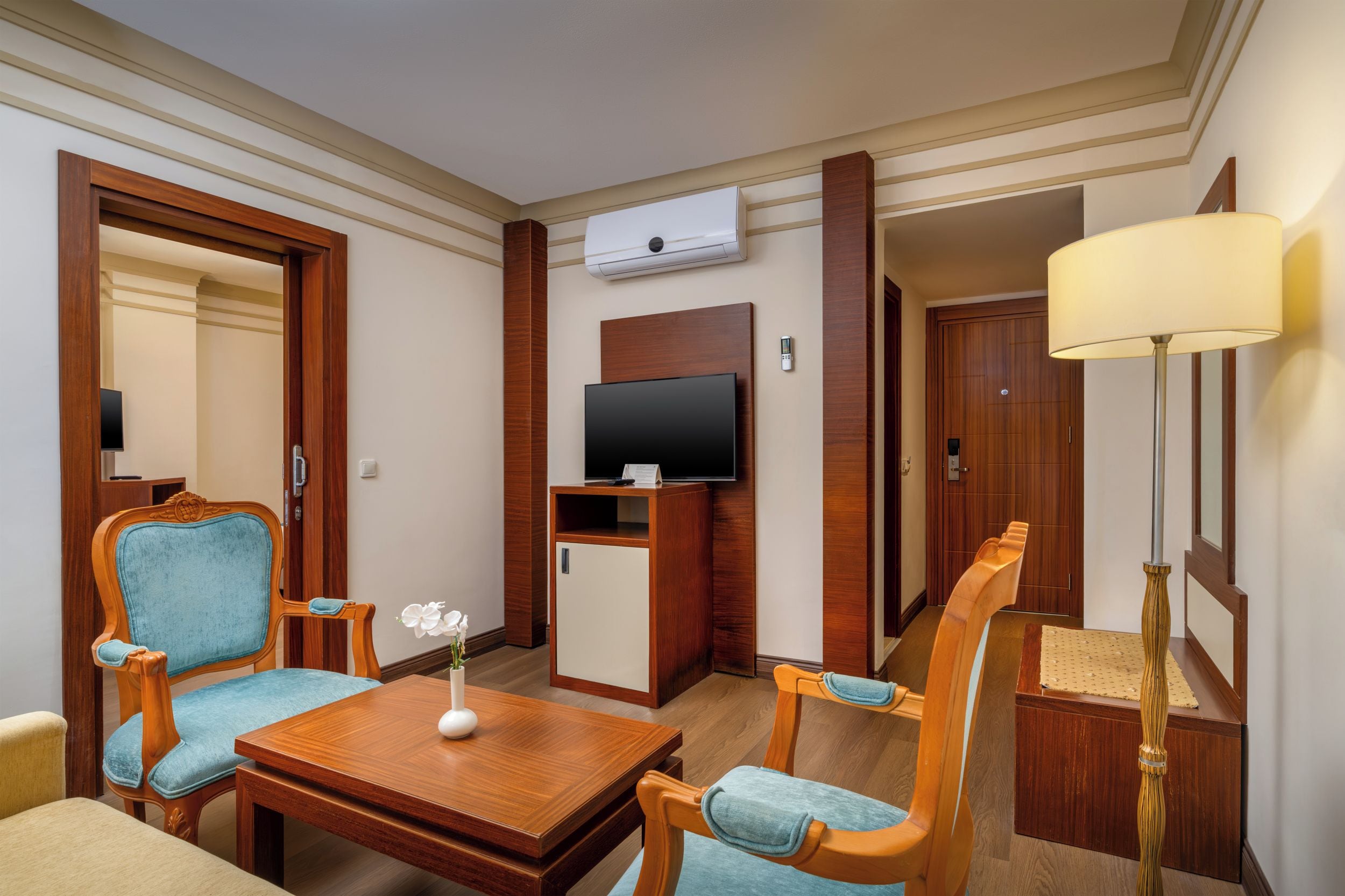 A hotel room with a wooden desk, two armchairs, a television, and an air conditioning unit.