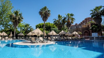 Un área de piscina con sillas de descanso y sombrillas, rodeada de palmeras y un edificio de piedra.