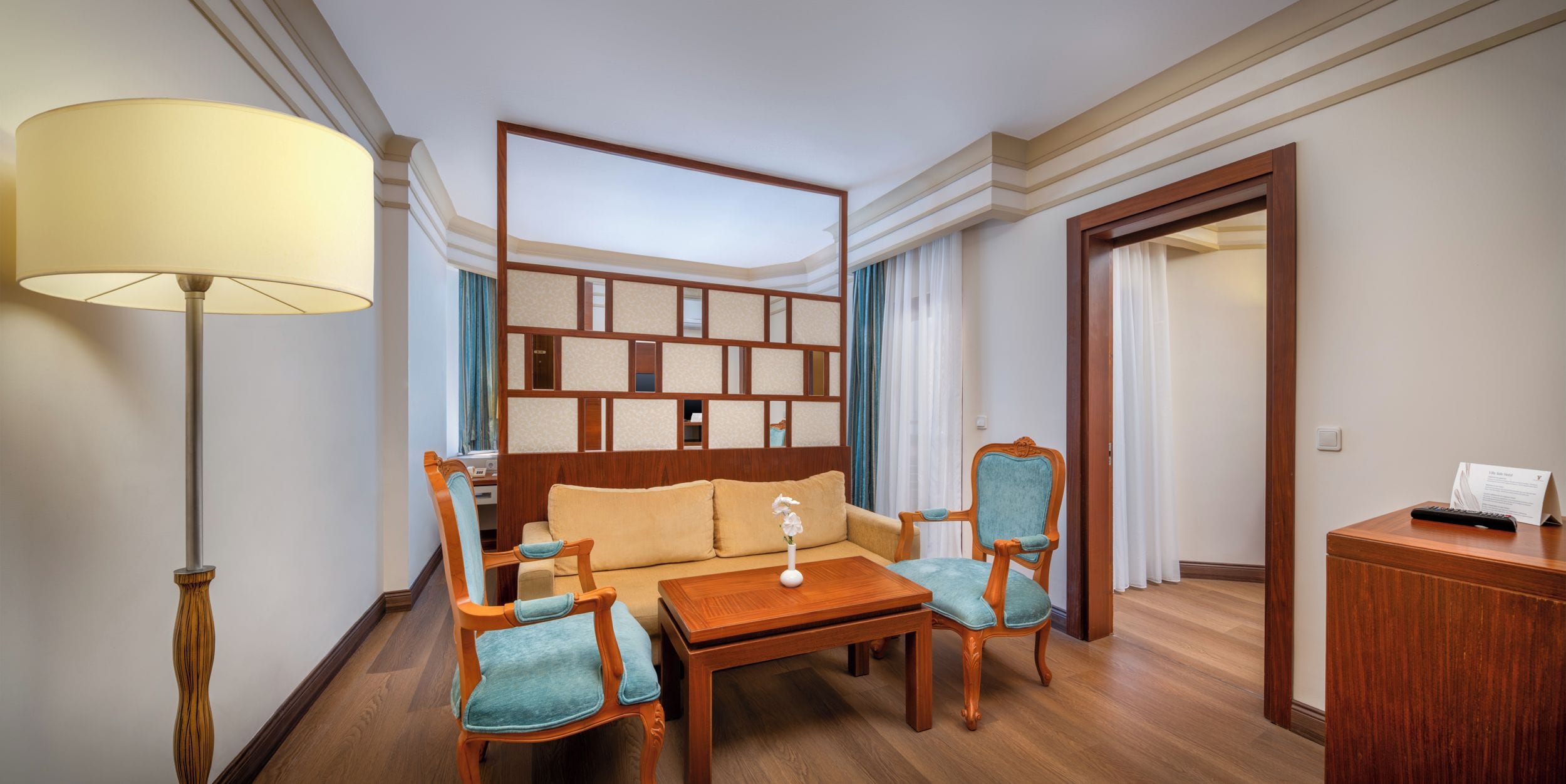 A hotel room with a wooden headboard, a sofa, two armchairs, a small table, a lamp, and a wooden cabinet.