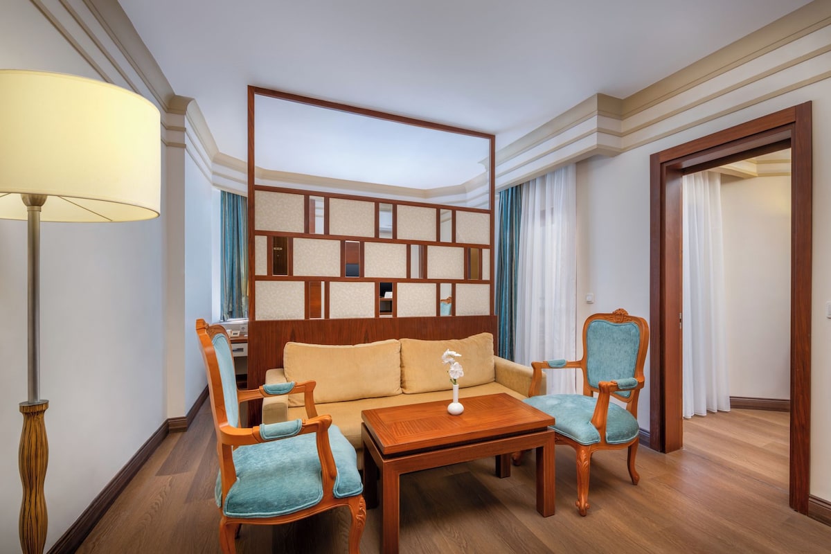 Une chambre d’hôtel avec une tête de lit en bois, un canapé, deux fauteuils, une petite table, une lampe et une armoire en bois.