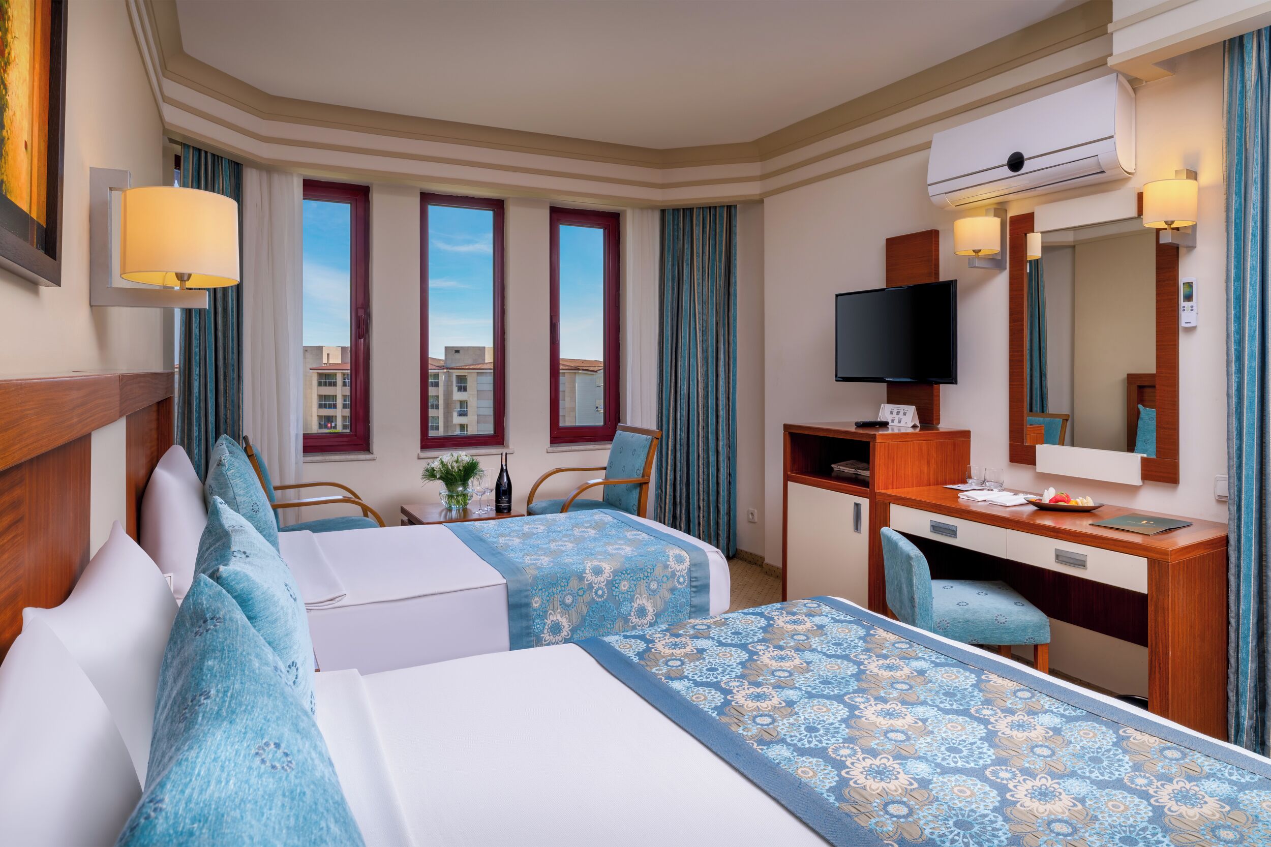 A hotel room with two beds, a desk, a television, and a large window with curtains.