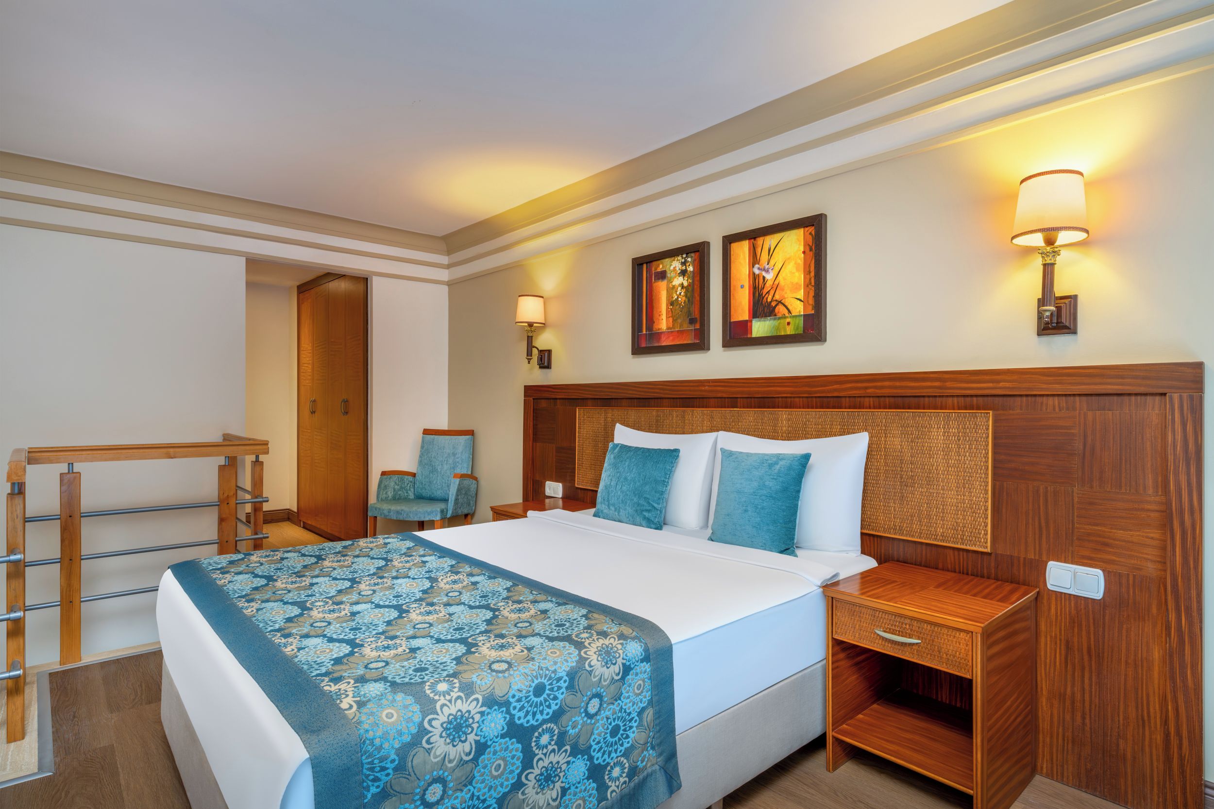 Une chambre d’hôtel avec un grand lit, une tête de lit en bois, deux lampes fixées au mur, une chaise, un bureau et une porte.