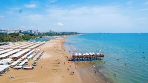 A beach with rows of sunbeds and umbrellas, a pier with umbrellas, and a clear blue sky. - Villa Side Hotel (Manavgat)
