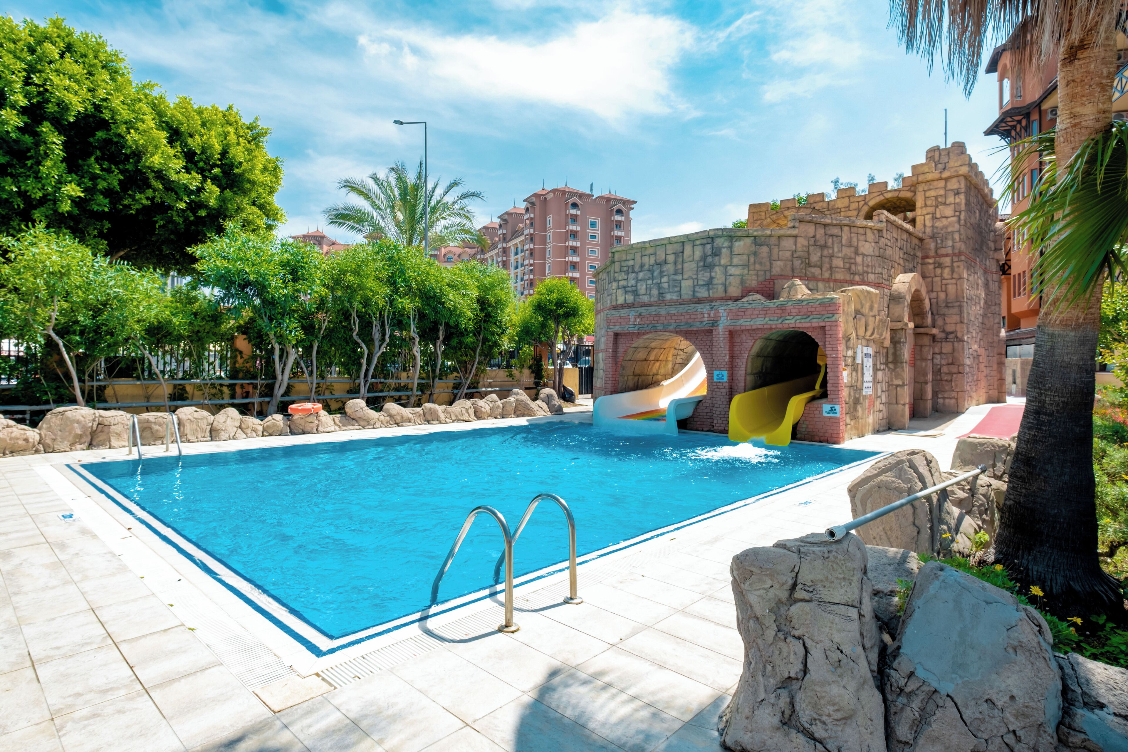 A swimming pool with a slide, surrounded by rocks and palm trees, with buildings in the background.