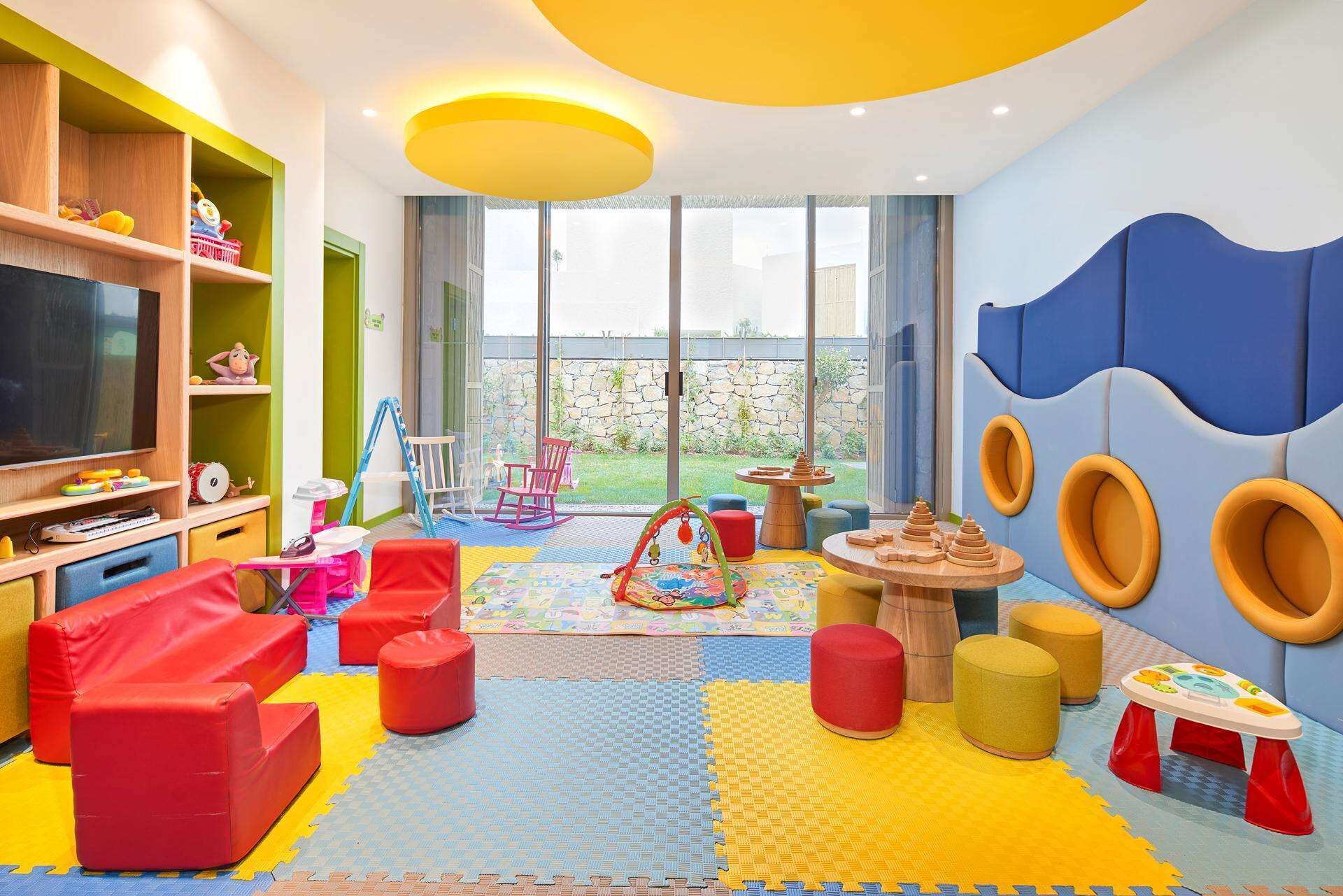 Une salle de jeux pour enfants aux couleurs vives, équipée d’un grand téléviseur, de tapis de jeu et de divers jouets.