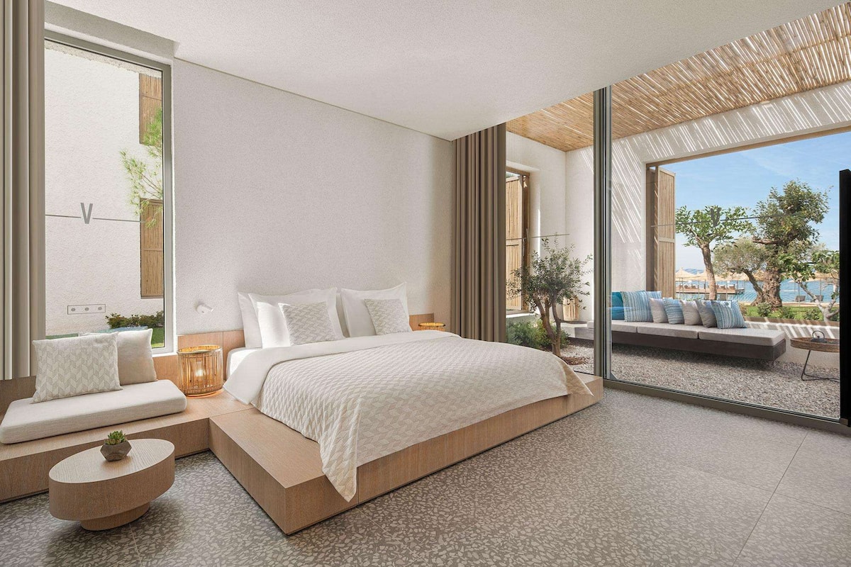 Ein modernes Schlafzimmer mit einem großen Bett, einer Sitzecke mit Kissen und Blick ins Freie.