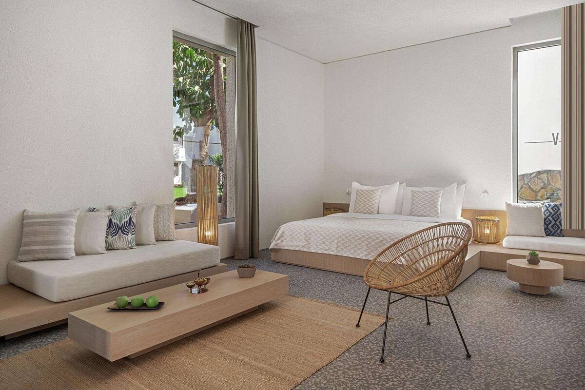Ein modernes Hotelzimmer mit einem Bett, einem Sofa, einem Korbstuhl, einem kleinen Tisch und einem großen Fenster mit Blick auf Grünflächen.