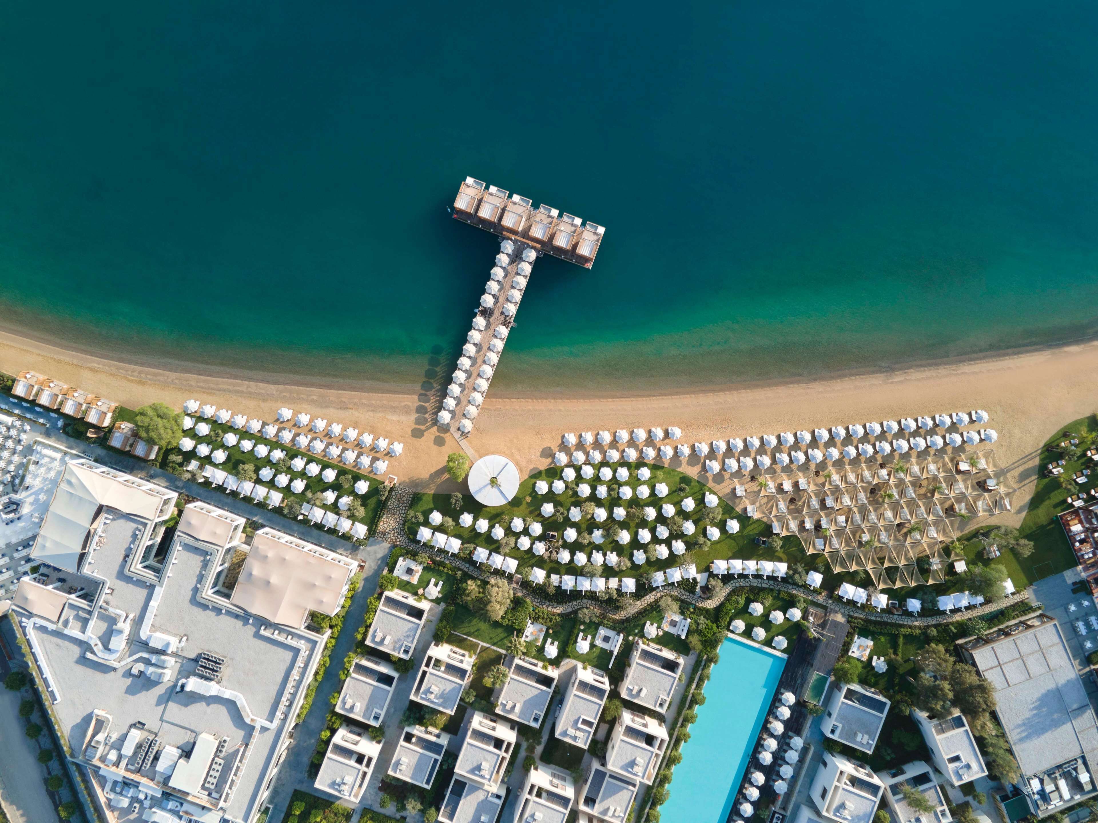 An aerial view of a coastal resort with a large T-shaped structure, multiple pools, and numerous beachside umbrellas.