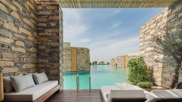 Una piscina de borde infinito con paredes de piedra, un muelle de madera, una zona de estar con cojines y sillas de descanso.