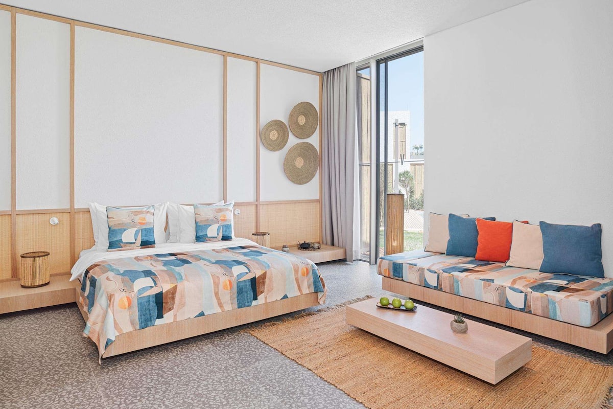 Ein modernes Hotelzimmer mit einem großen Bett, einer Couch, einem Couchtisch und Blick ins Freie.