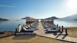 Muelles de madera que conducen a sillones de playa con sombrillas, con vista a aguas tranquilas y colinas lejanas.