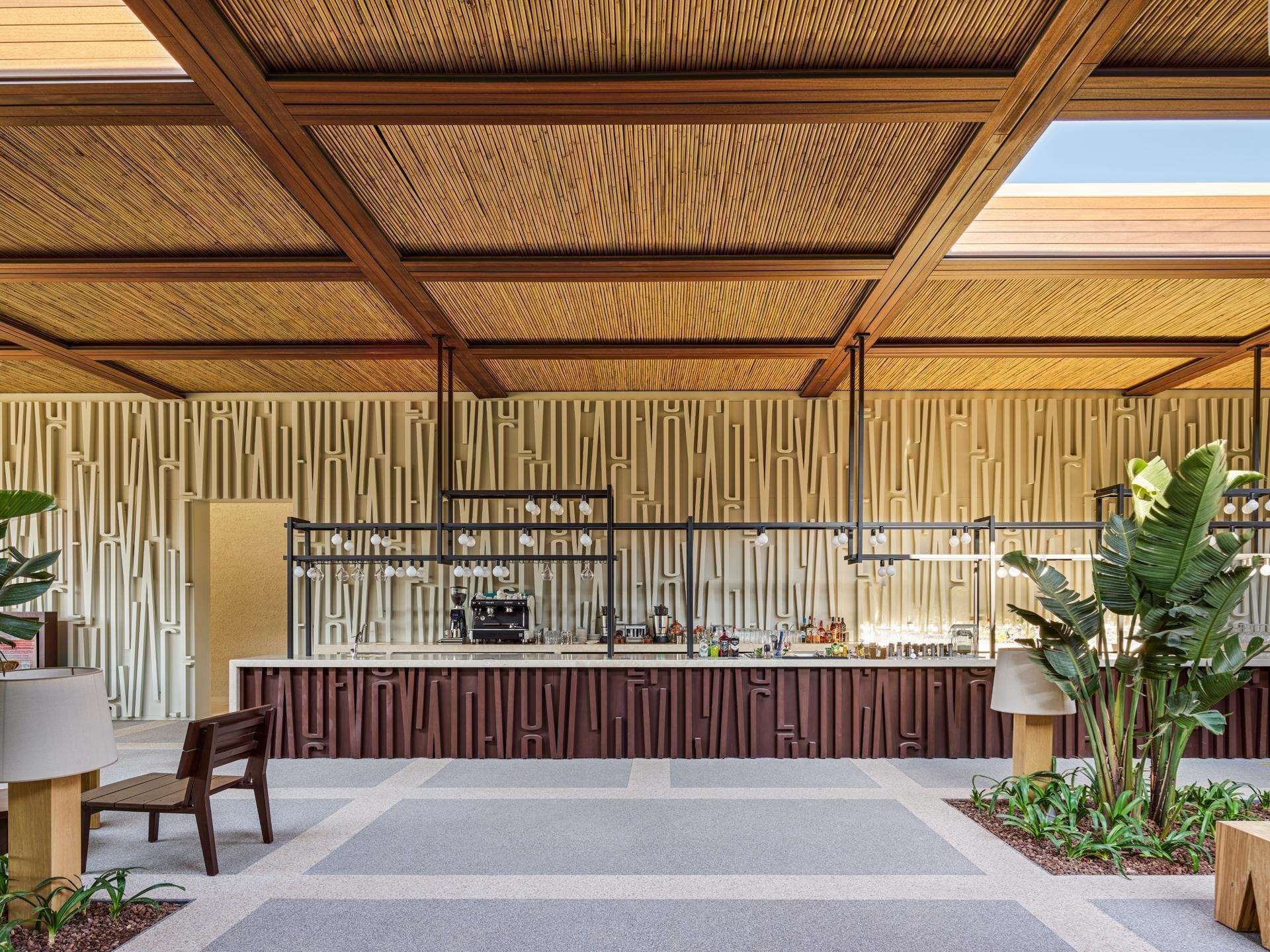 Un área de bar moderna al aire libre con techo de madera, mostrador con equipo de bar, asientos y plantas.
