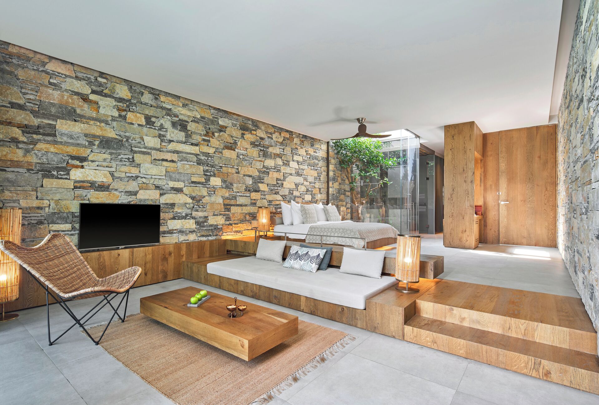 A modern living room with a stone wall, a flat-screen TV, a sofa, a coffee table, and a staircase leading to another room.