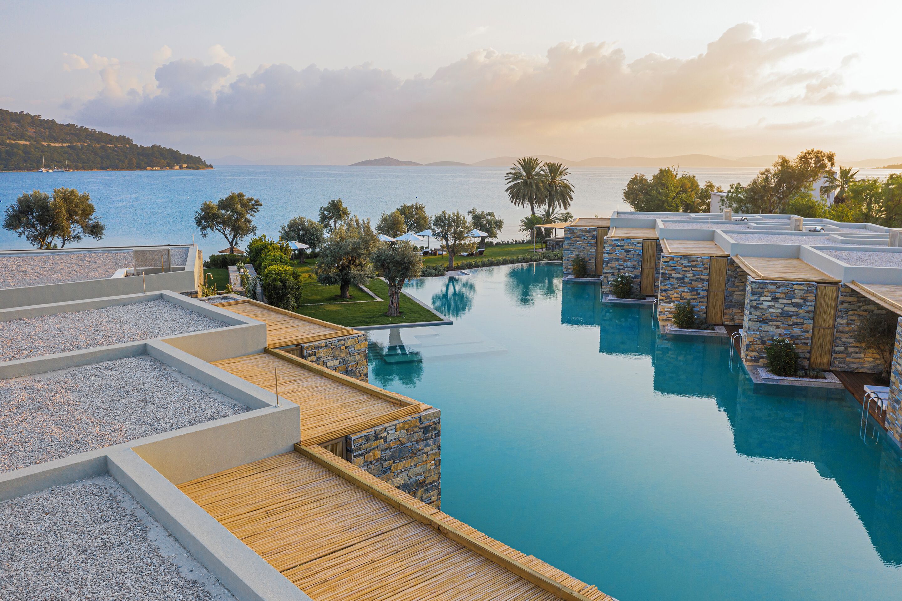 A pool area with multiple swimming pools, wooden deck, stone walls, and a view of the sea.