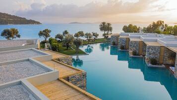 A pool area with multiple swimming pools, wooden deck, stone walls, and a view of the sea.