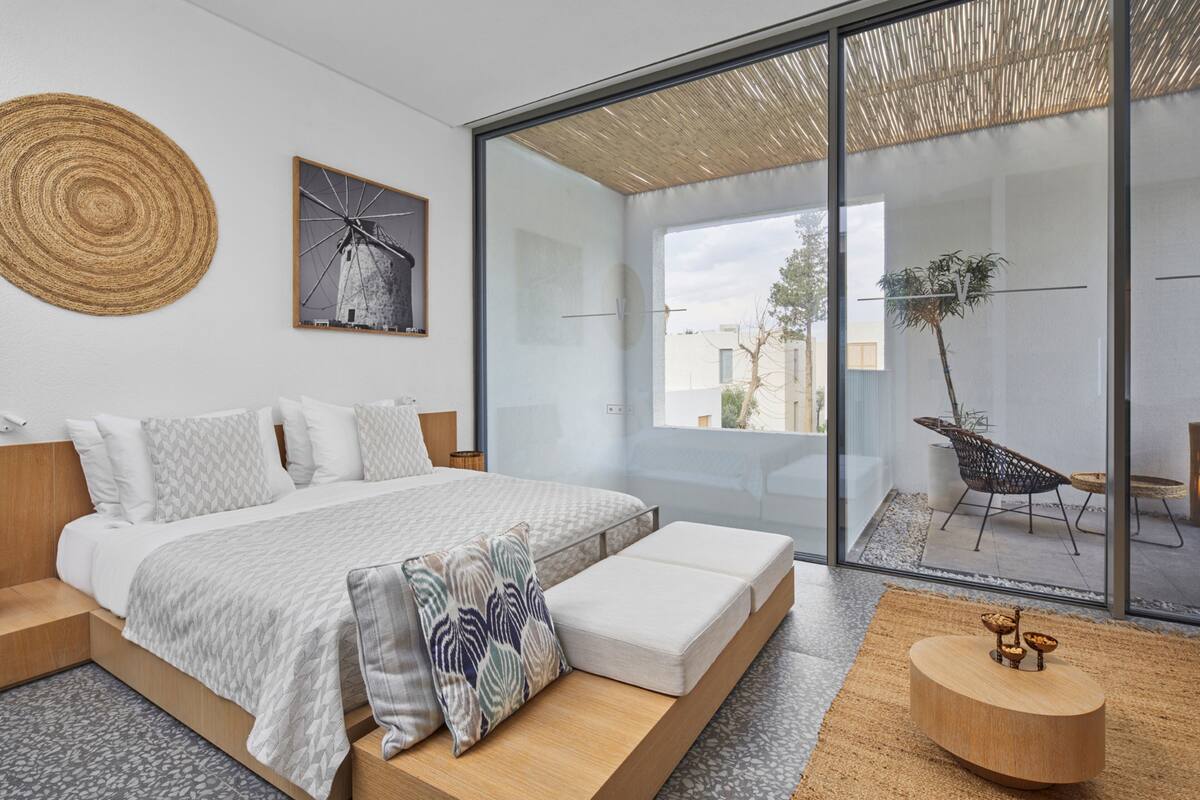 Ein modernes Schlafzimmer mit einem großen Bett, Blick ins Freie und einer kleinen Sitzecke mit einer Pflanze.