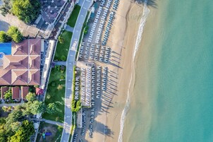 Vista aérea de un hotel frente a la playa, con hileras de sombrillas, un estacionamiento y un edificio con un diseño distintivo en el techo.