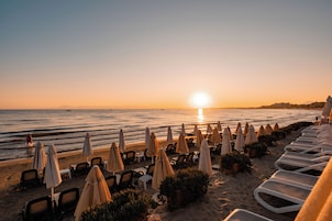 Un hotel frente a la playa con filas de tumbonas y sombrillas, vista despejada del océano y un atardecer de fondo.