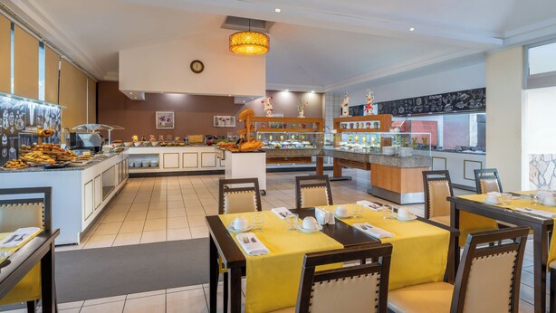 Un comedor de hotel con mostradores de buffet, mesas preparadas para el desayuno y una cocina al fondo.