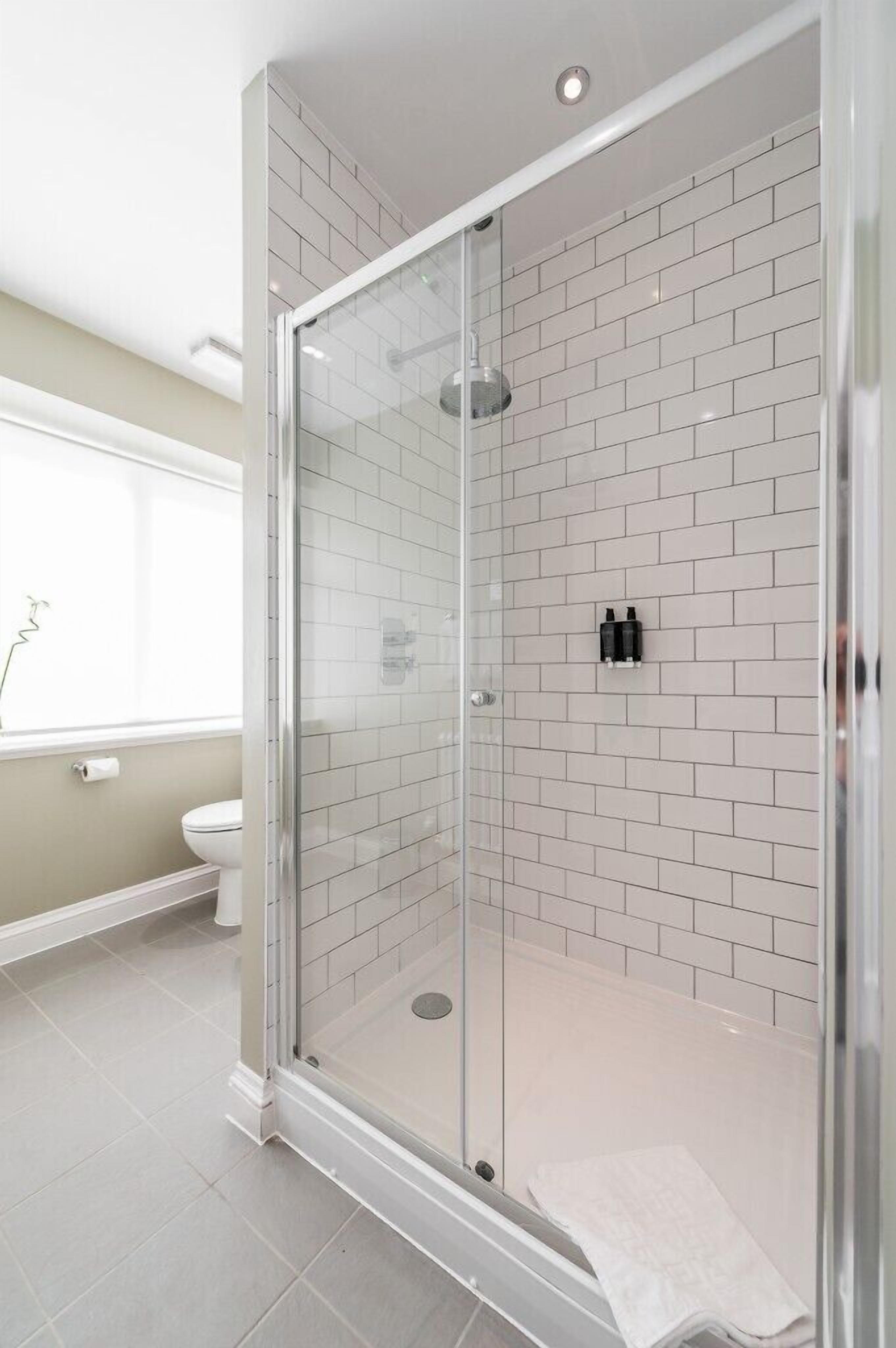 A modern bathroom with a glass-enclosed shower, white tiled walls, and a toilet visible in the background.