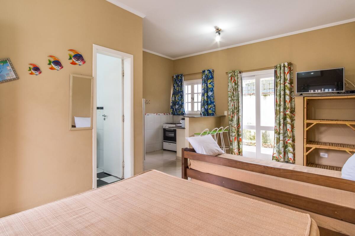 A bedroom with a wooden bed, a TV on a shelf, and a kitchen area with a stove and oven.