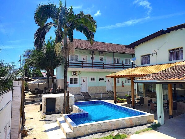 A swimming pool with a stone border, a covered patio area, and a two-story building with a tiled roof. - Recanto Maranduba (Ubatuba)