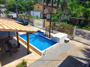 A swimming pool with a pergola and outdoor seating, surrounded by a fenced yard with houses and trees. - Recanto Maranduba (Ubatuba)