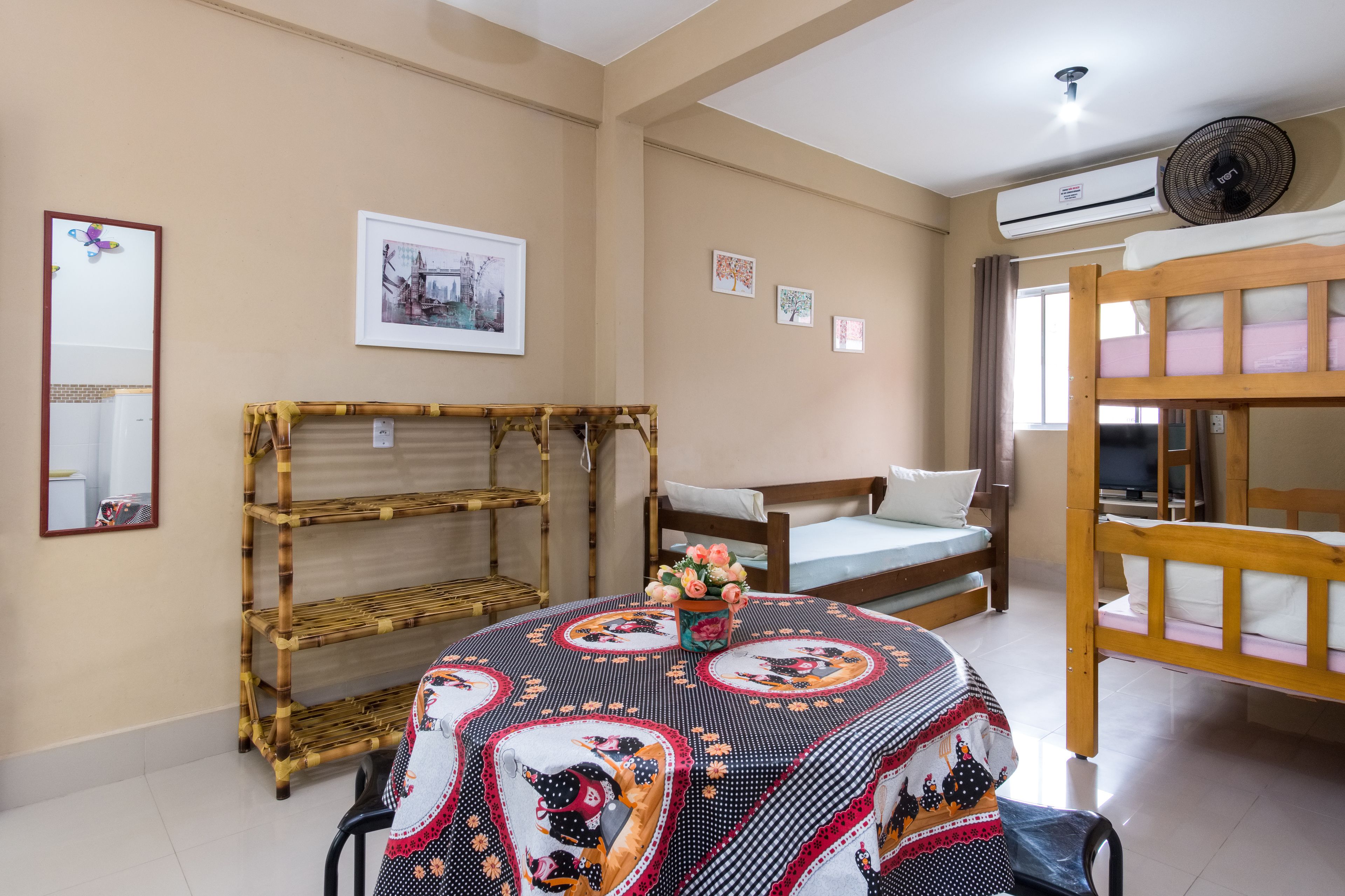 A room with a bunk bed, a single bed, a bamboo shelf unit, and a table with a patterned tablecloth.