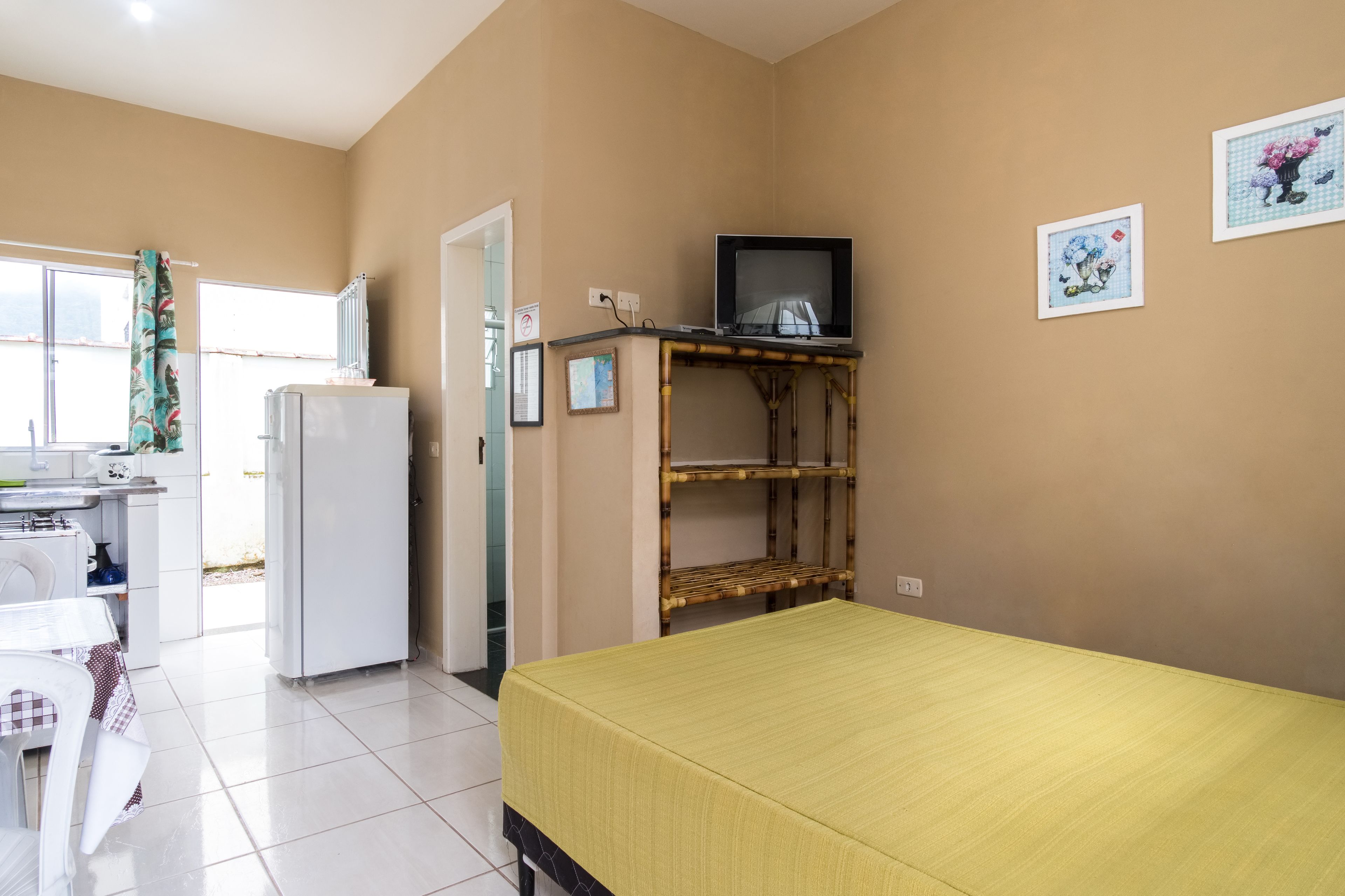 Habitación compacta con cama, una kitchenette pequeña, un televisor sobre una repisa de bambú y una ventana con cortinas.