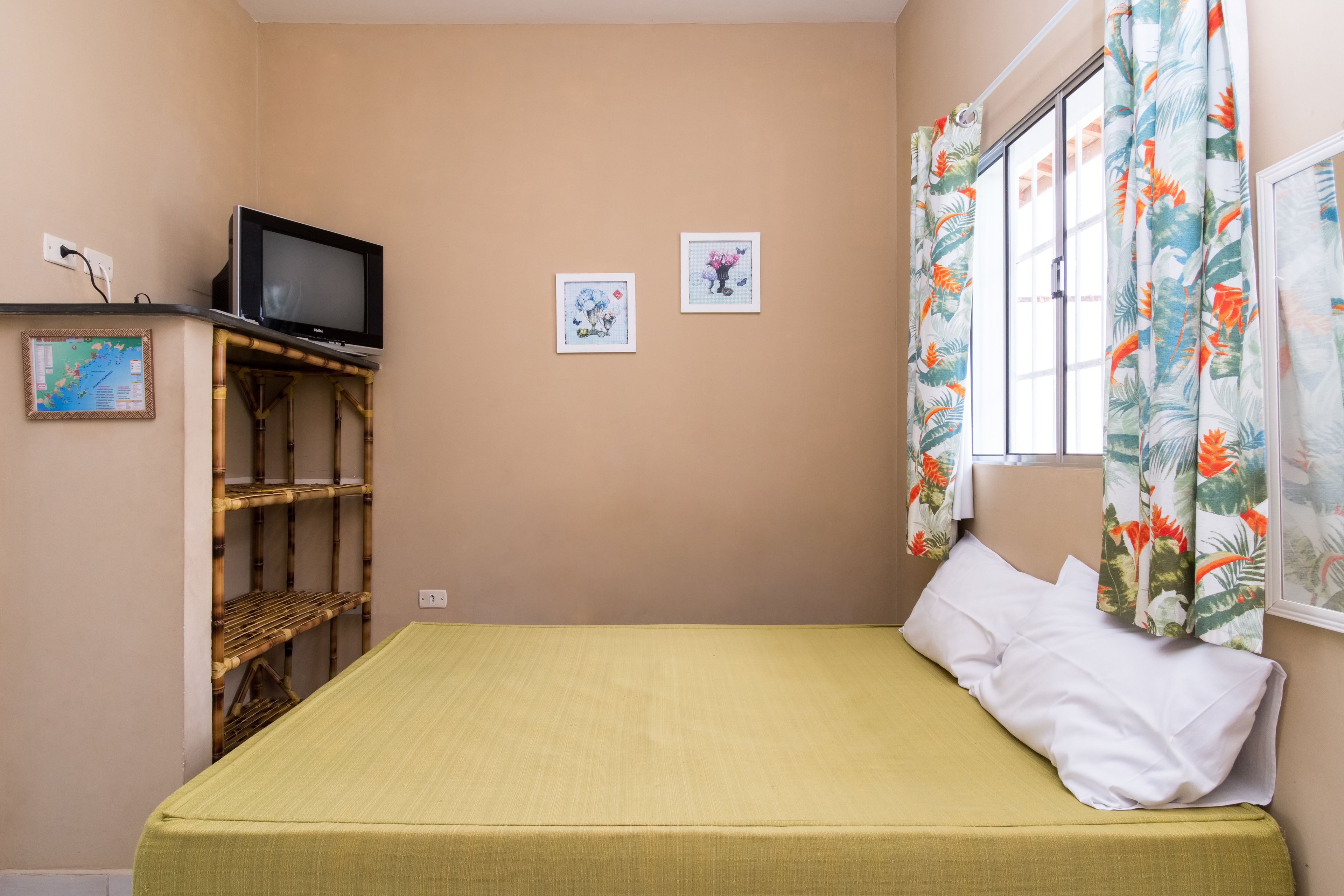 Una habitación pequeña con una cama individual, una colcha verde, un televisor sobre una estantería de bambú y dos cuadros en la pared.