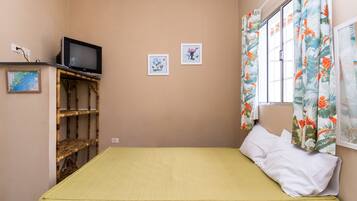 Una habitación pequeña con una cama individual, una colcha verde, un televisor sobre una estantería de bambú y dos cuadros en la pared.