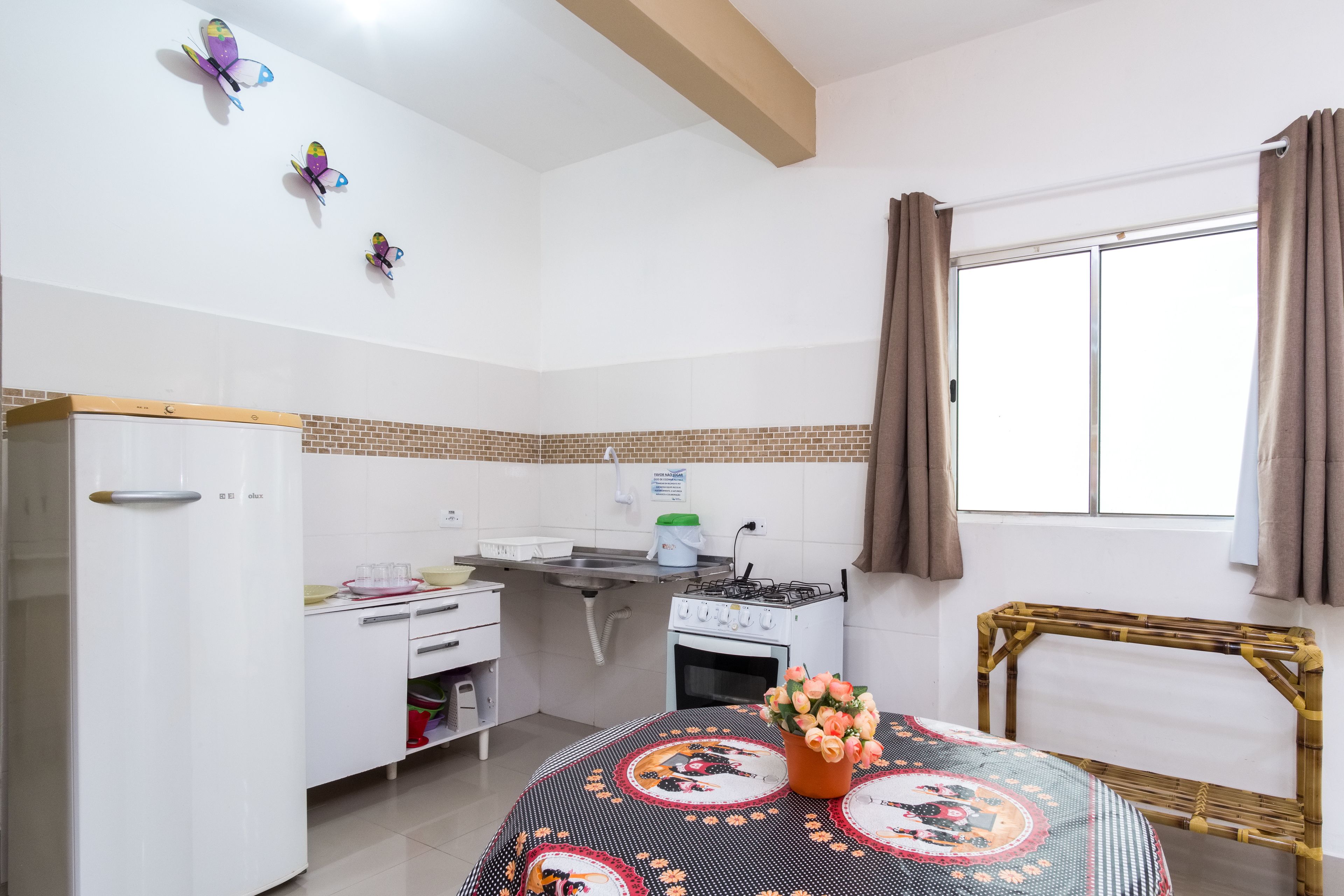 A kitchen with a white refrigerator, a stove, and a sink. There is a window with curtains and a decorative wall hanging.