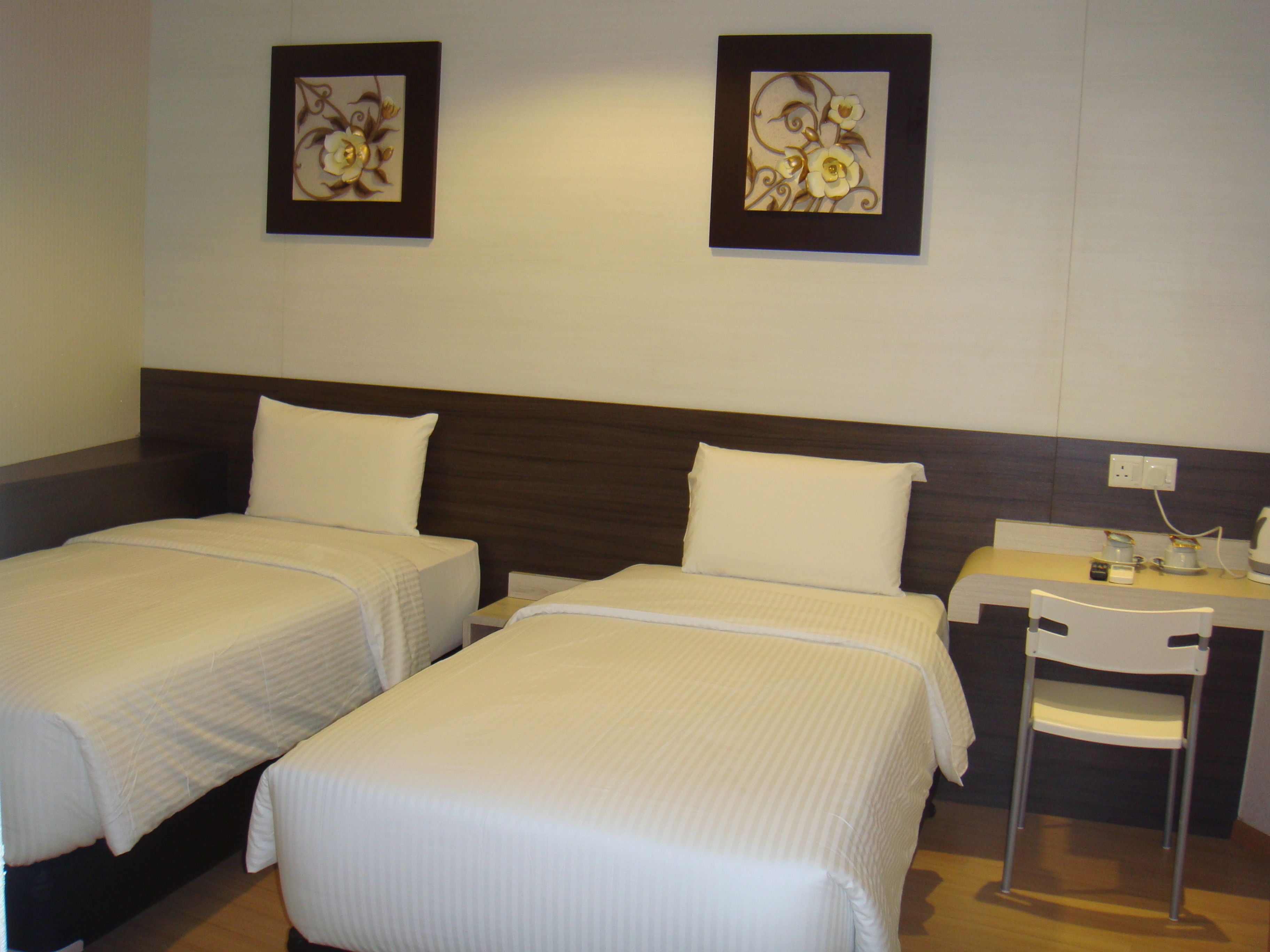 A hotel room with two single beds, a small desk with a chair, and two framed floral artworks on the wall.