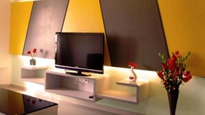 A modern living room with a flat-screen TV on a white stand, a small white shelf with a vase of red flowers, and a decorative vase with red flowers on the right. - Duta Hotel & Residence (Kuala Lumpur)