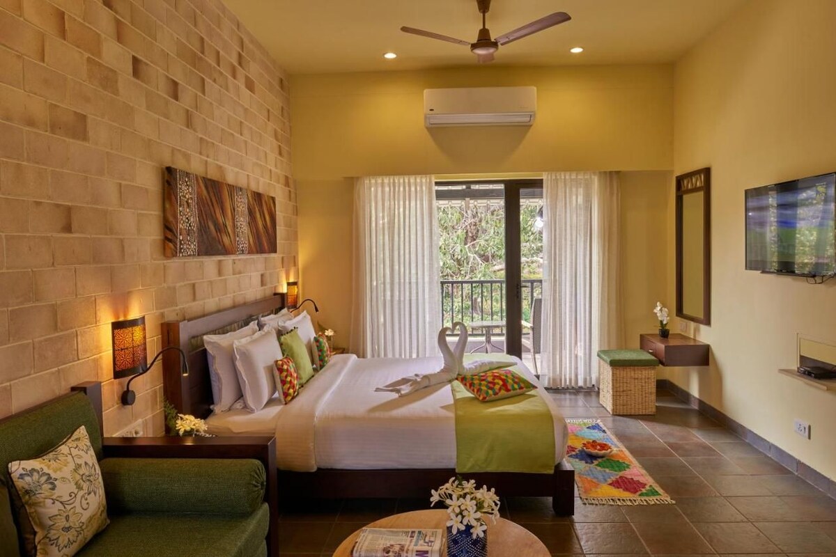 A hotel room with a bed, a green sofa, a small table with flowers, a TV, and a balcony with curtains.