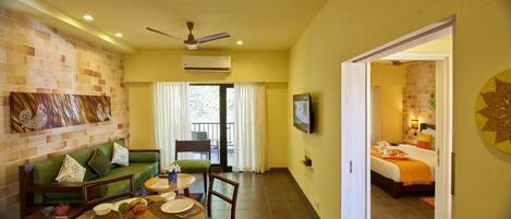 Una habitación de hotel con un sofá verde, una mesa de comedor con platos y frutas, un televisor y un balcón con cortinas.