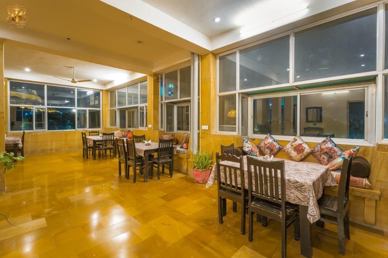 Uma sala de jantar ampla, com pé direito alto e grandes janelas, com mesas e cadeiras de madeira e uma planta em vaso.