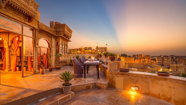 A rooftop terrace with a dining area, potted plants, and a view of the cityscape at sunset. - Hotel Garh Jaisal Haveli (Jaisalmer)