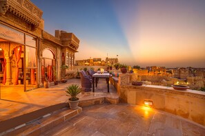 A rooftop terrace with a dining area, potted plants, and a view of the cityscape at sunset. - Hotel Garh Jaisal Haveli (Jaisalmer)