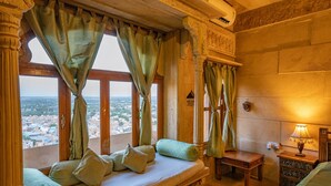 A room with a bed, a small table, a lamp, and a view of a cityscape through large windows with curtains. - Hotel Garh Jaisal Haveli (Jaisalmer)