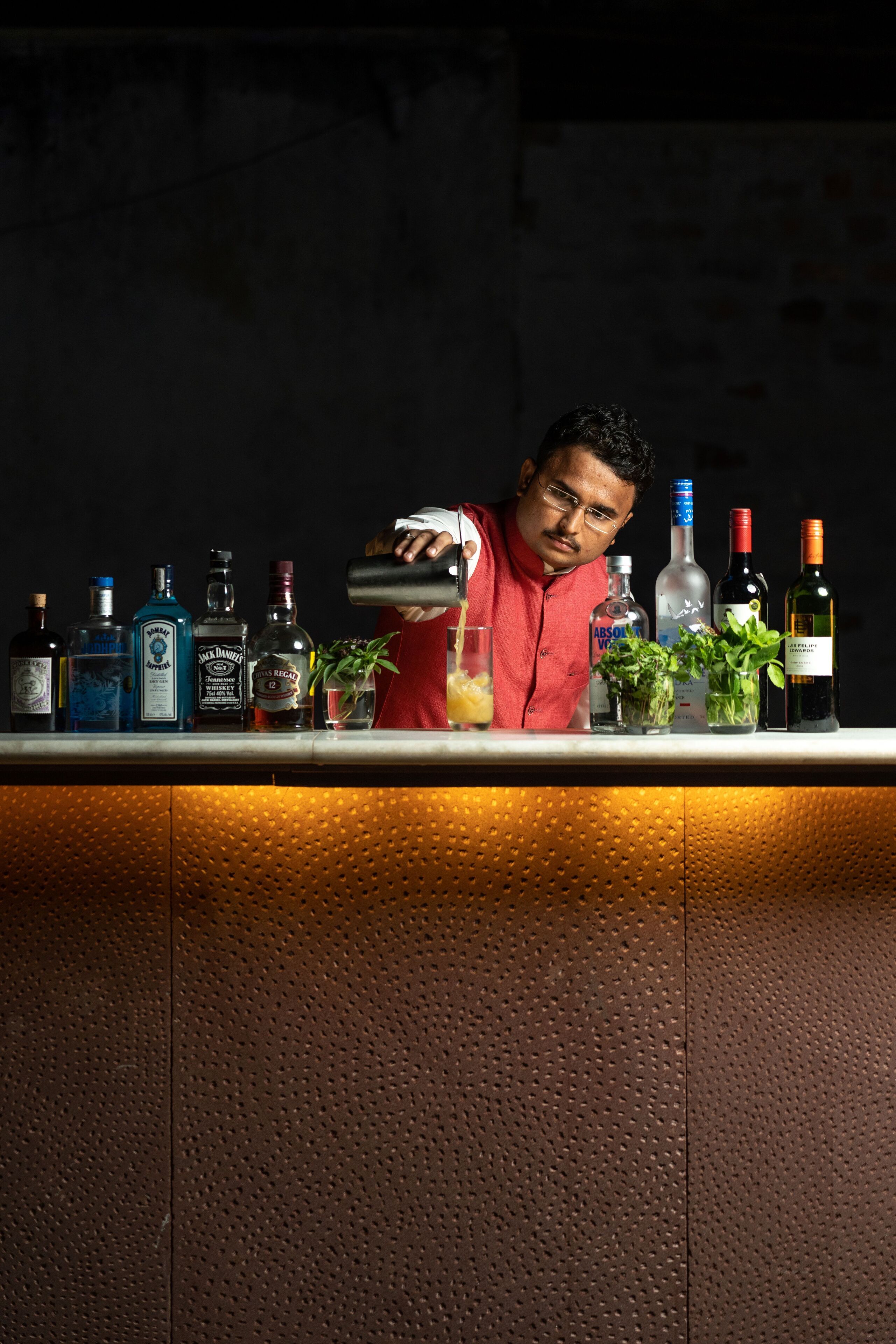 A bar with a bartender preparing a drink, various bottles of alcohol, and small potted plants on the counter.