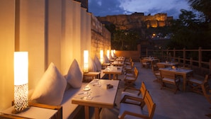 Un área de comedor al aire libre con mesas y sillas, iluminada por lámparas fijadas a la pared.