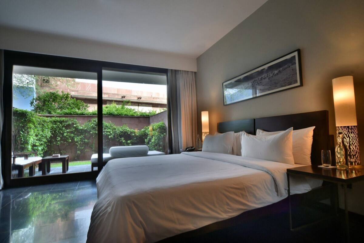 A hotel room with a large bed, a desk, a chair, a nightstand, a lamp, and a view of a courtyard with greenery.