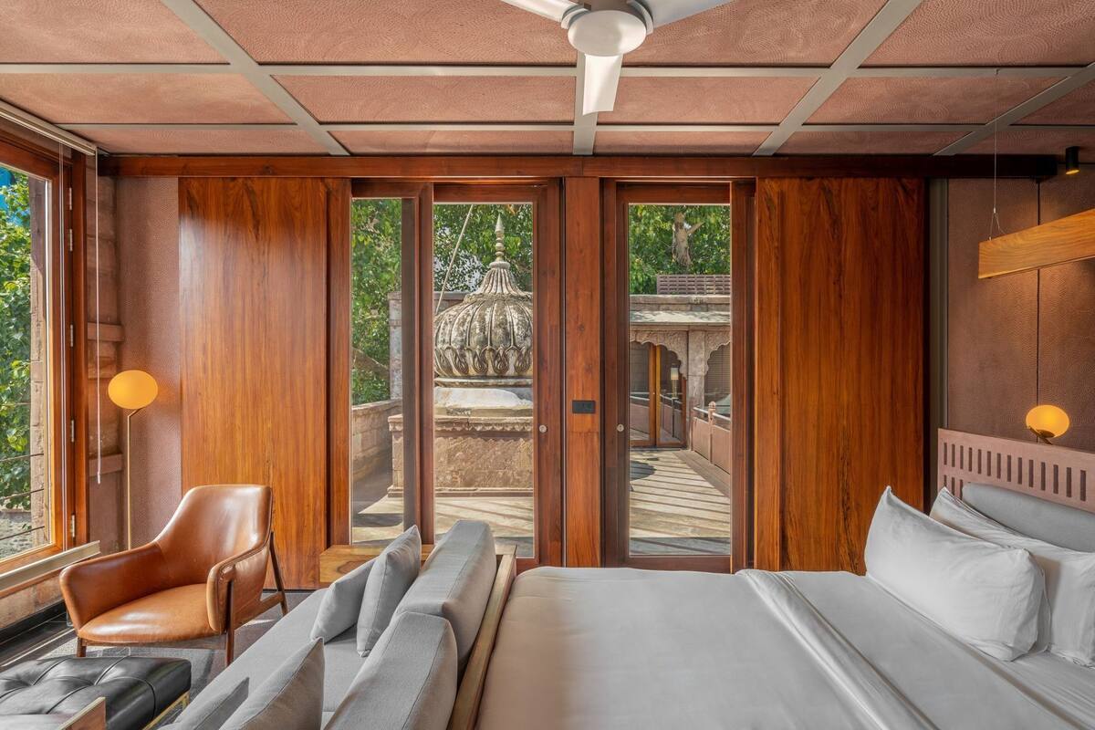 A room with a bed, a chair, a sofa, a ceiling fan, and a view of an outdoor area with a stone structure.