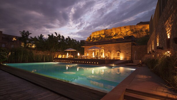 A swimming pool with a wooden deck, a stone building with a rock formation, and outdoor seating with umbrellas. - RAAS Jodhpur (Jodhpur)