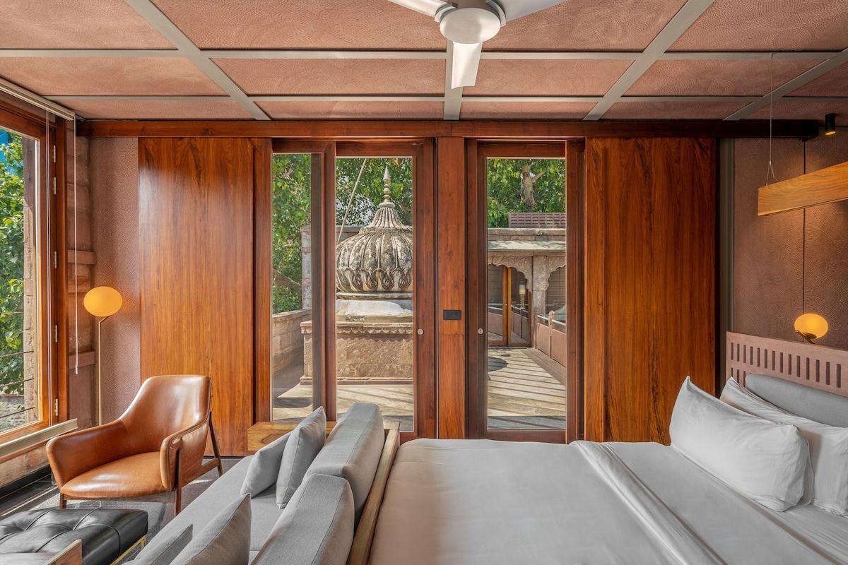 A room with a bed, a chair, a sofa, a ceiling fan, and a view of an outdoor area with a stone structure.